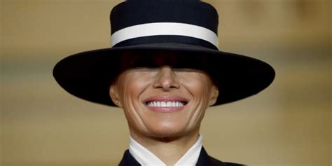 a close-up image of a woman wearing a large black hat with a white band that obscures her eyes, she is smiling broadly. The…