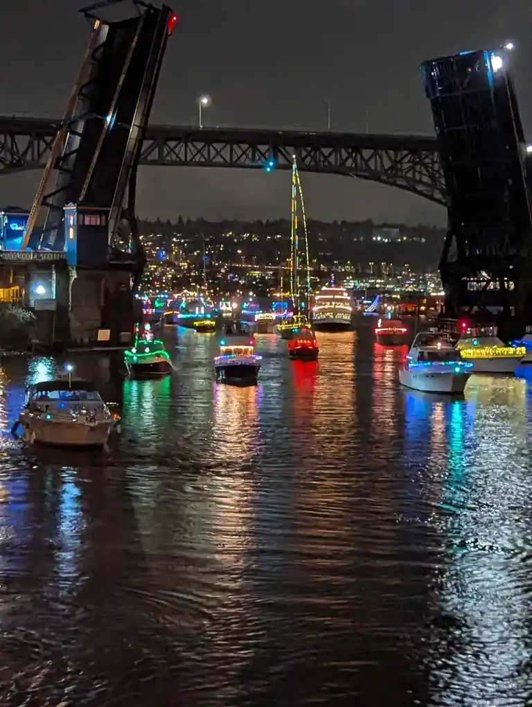 A nighttime photo shows numerous boats decorated with colorful lights navigating under a raised drawbridge. The water…