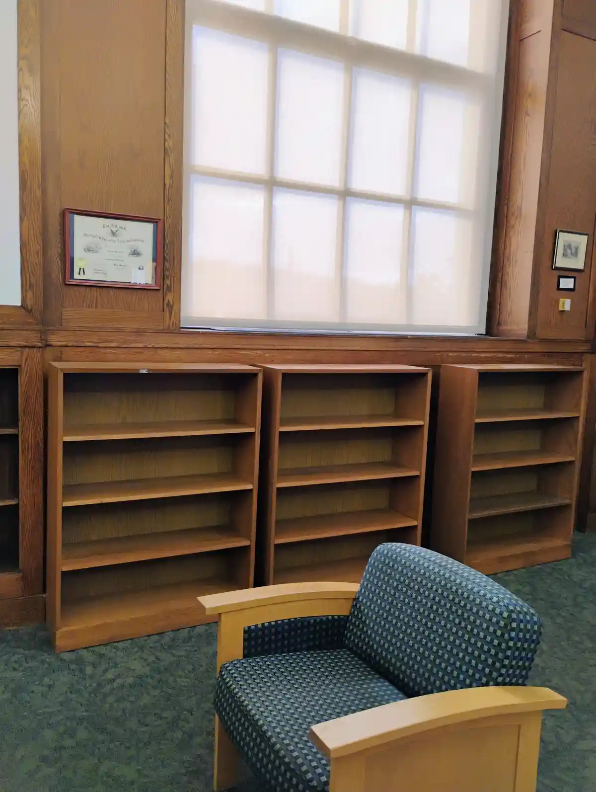 An empty wooden bookshelf sits in a library next to a window with blinds, with a patterned armchair in the foreground. The…