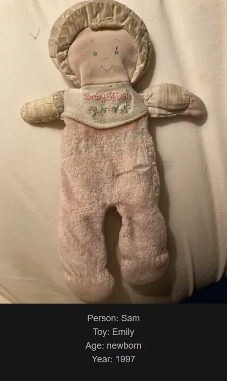 A faded, pink, vintage baby doll with a stitched face and bonnet lies on a white fabric surface. Overlaid text identifies…