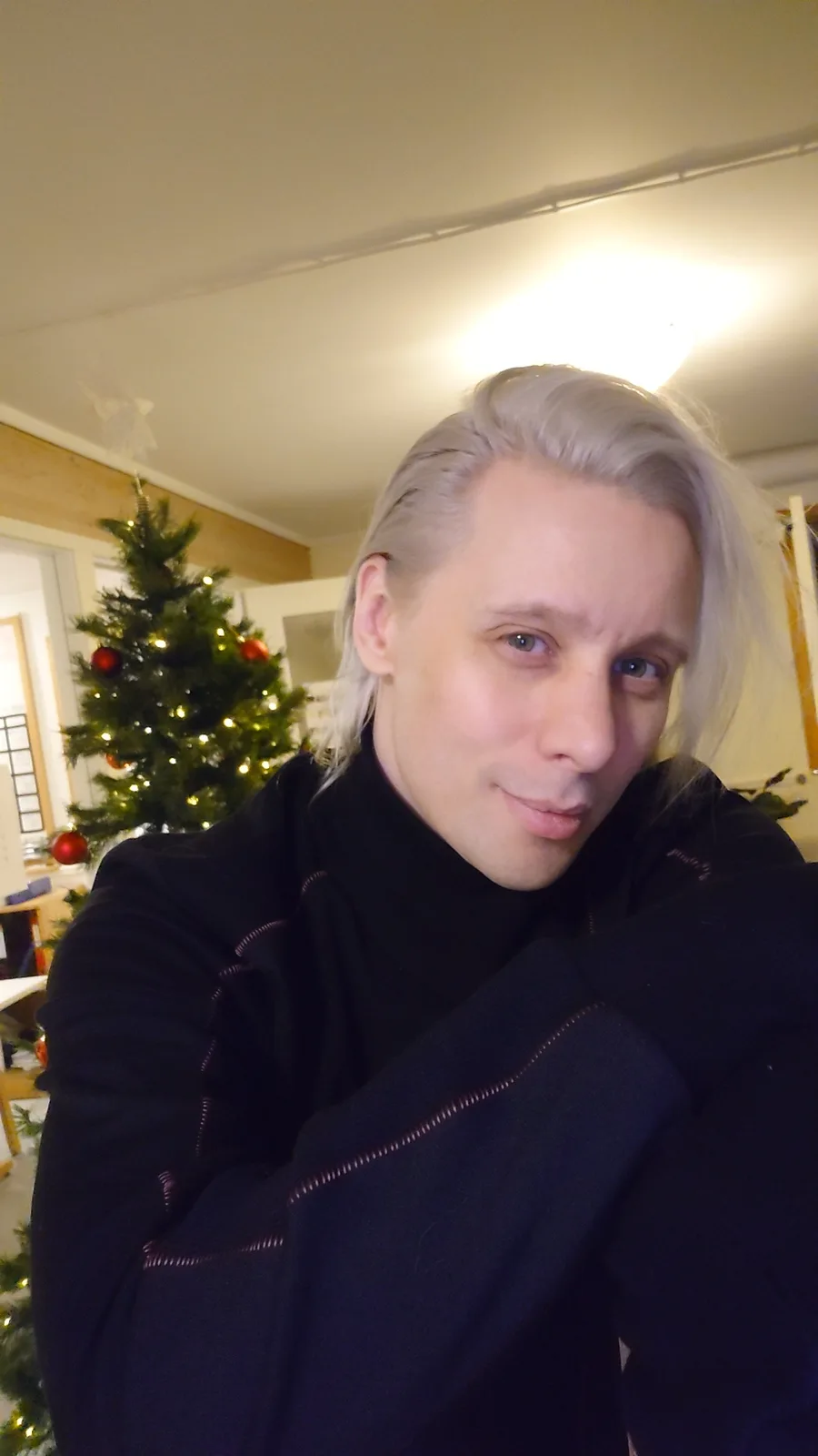 A person with long, platinum blonde hair and a black turtleneck sweater poses in front of a decorated Christmas tree with…