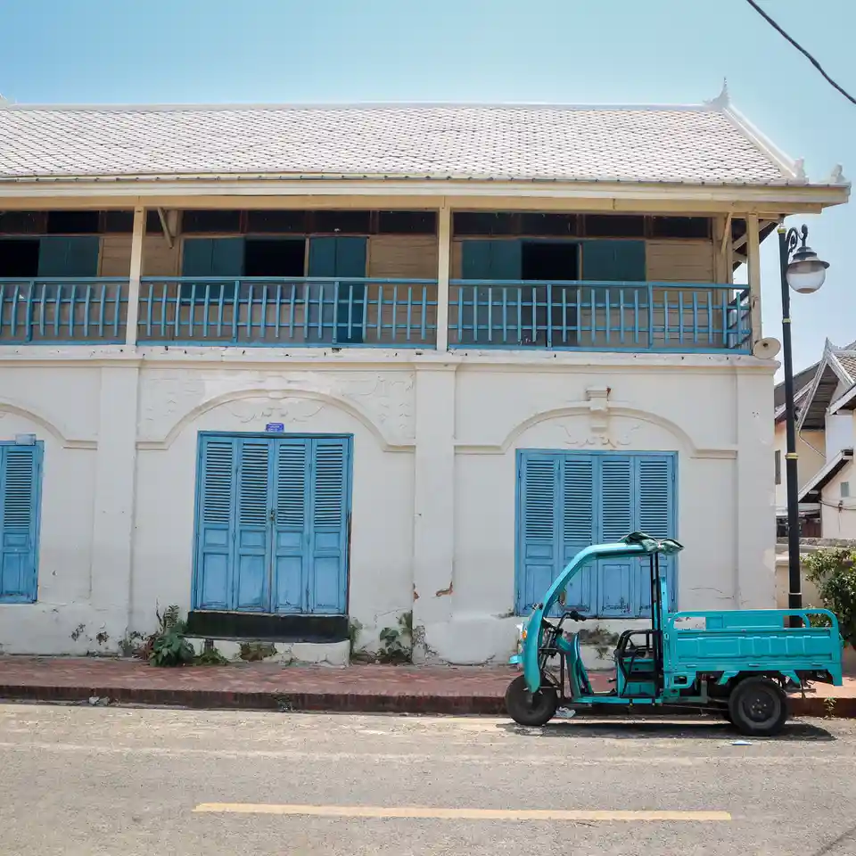 A turquoise three-wheeled cargo tricycle is parked on a street in front of a white building with blue shutters and a…