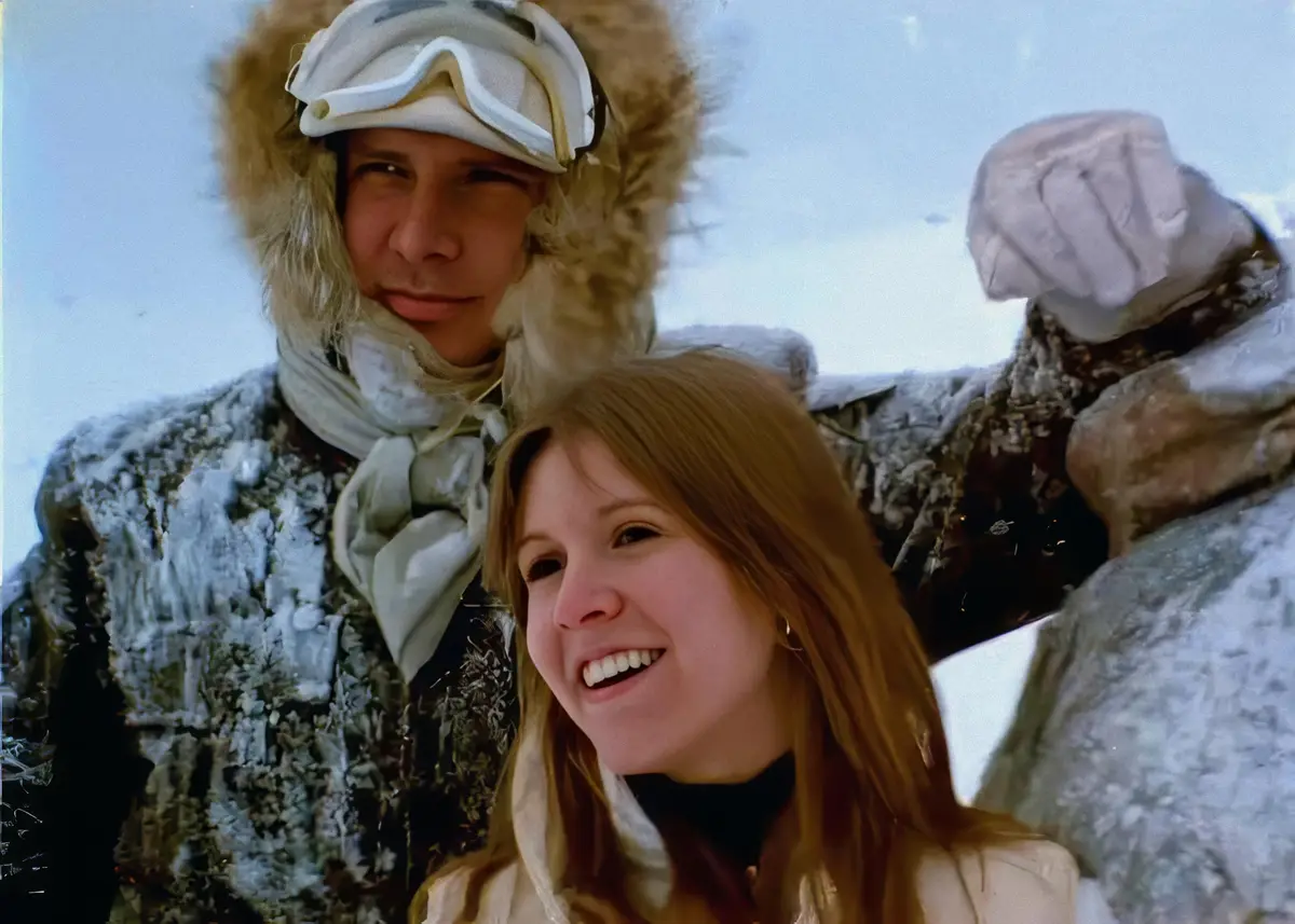 A man and a woman are in a snowy landscape, both dressed in heavy winter gear. The man wears a fur-lined hood and goggles…