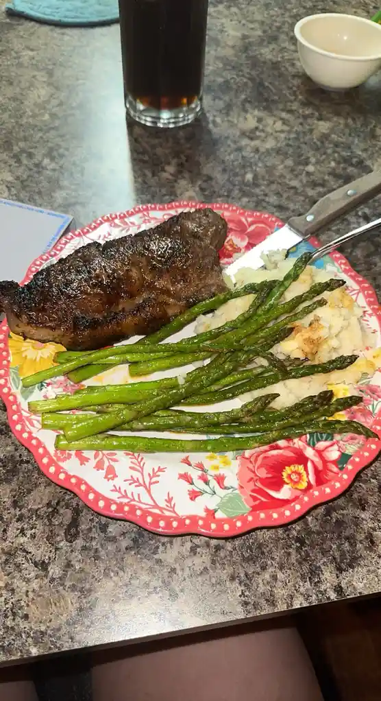A plate of grilled steak, roasted asparagus, and mashed potatoes sits on a floral-patterned plate with a knife resting on…