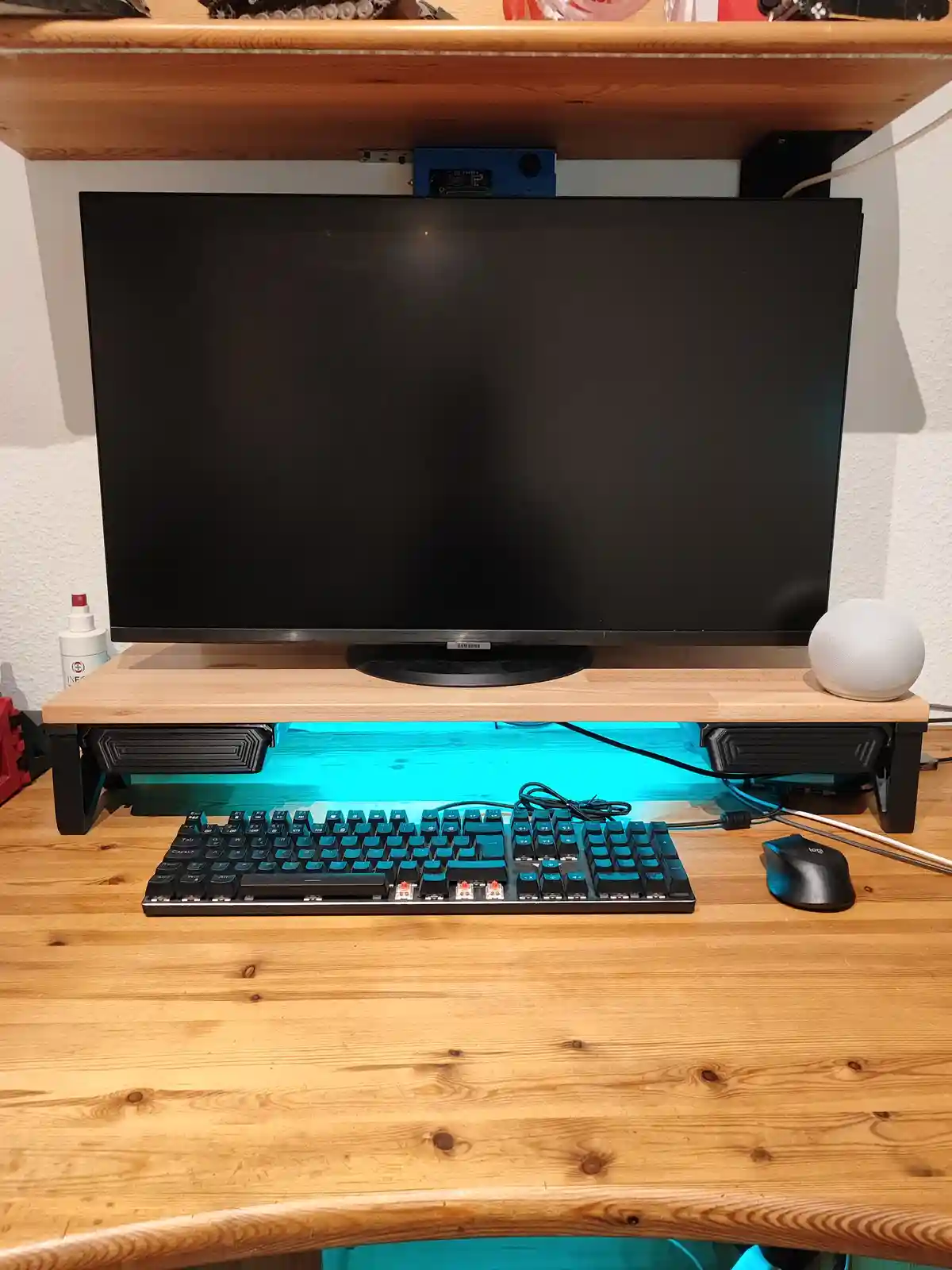 A wooden desk setup with a black Samsung monitor on a riser, a mechanical keyboard with exposed red switches in front, and a…