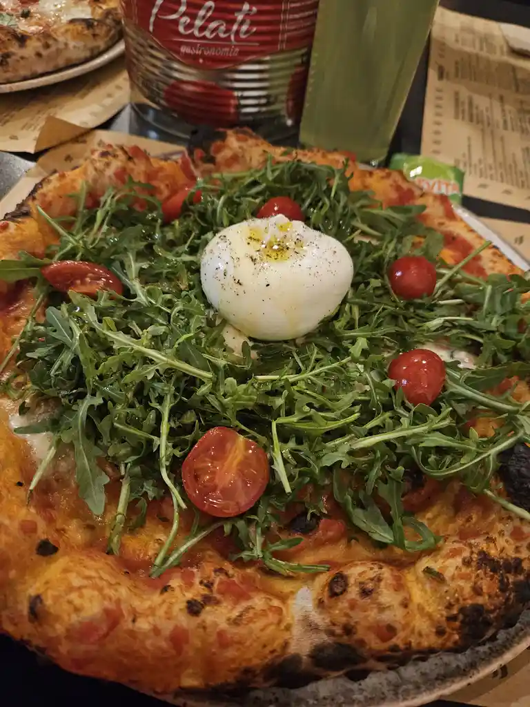 A close-up photo of a pizza topped with arugula, halved cherry tomatoes, and a large, centered ball of burrata cheese…
