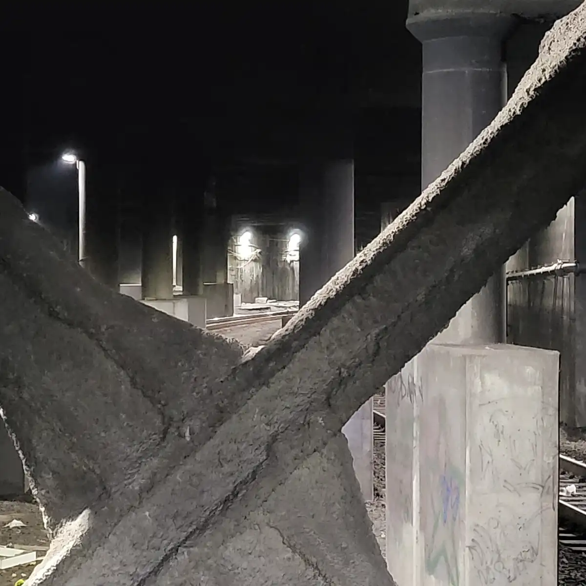 A nighttime photo of a dark, abandoned underpass or bridge structure with concrete pillars and beams, illuminated by sparse…