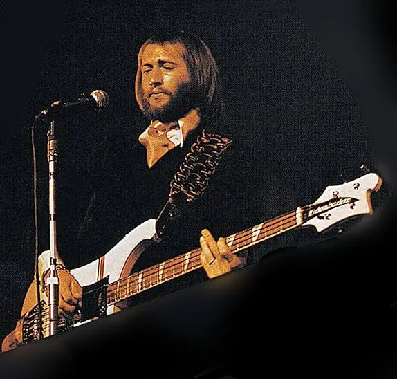 a man with long hair and a beard, wearing a dark shirt and bowtie, plays a white bass guitar on stage under dim lighting…