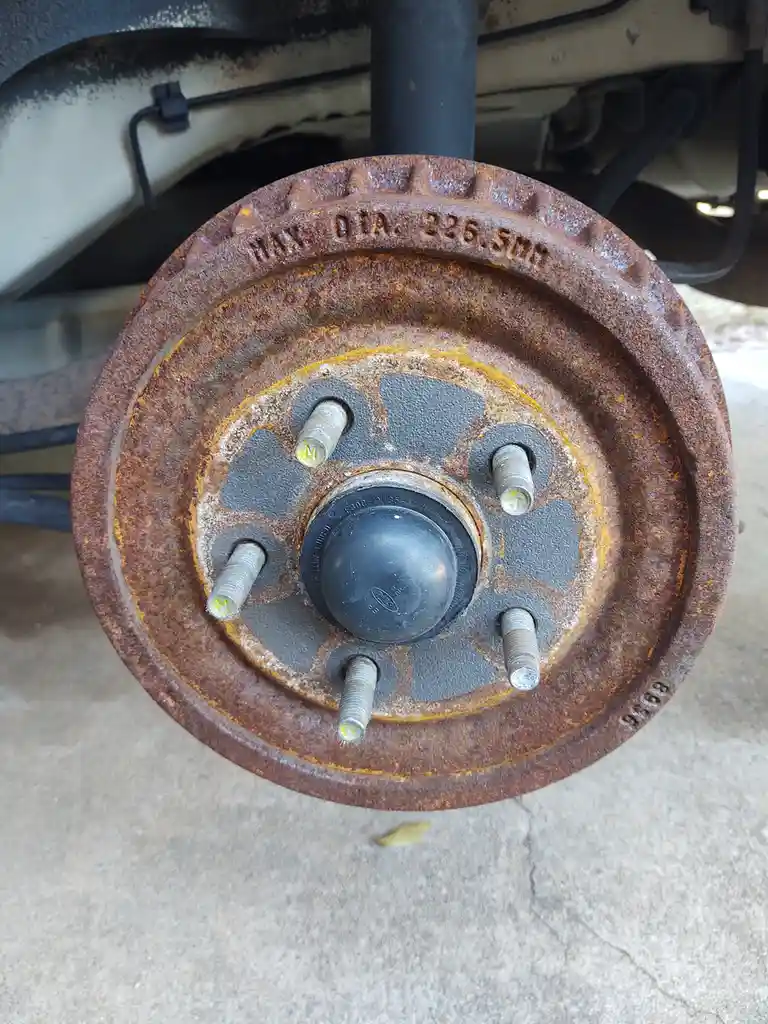 A close-up photo shows a rusty, circular metal wheel hub with five bolts protruding from its center, set against a concrete…