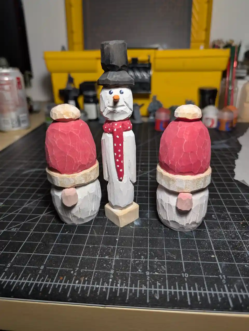 Three hand-carved wooden figurines sit on a gridded cutting mat: a central snowman with a black top hat and red scarf…