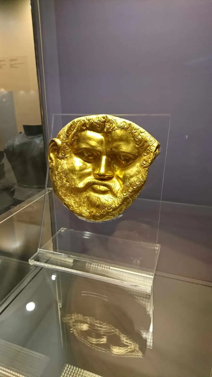 A golden, ancient Greek-style death mask with a beard and curly hair is displayed on a clear acrylic stand inside a glass…