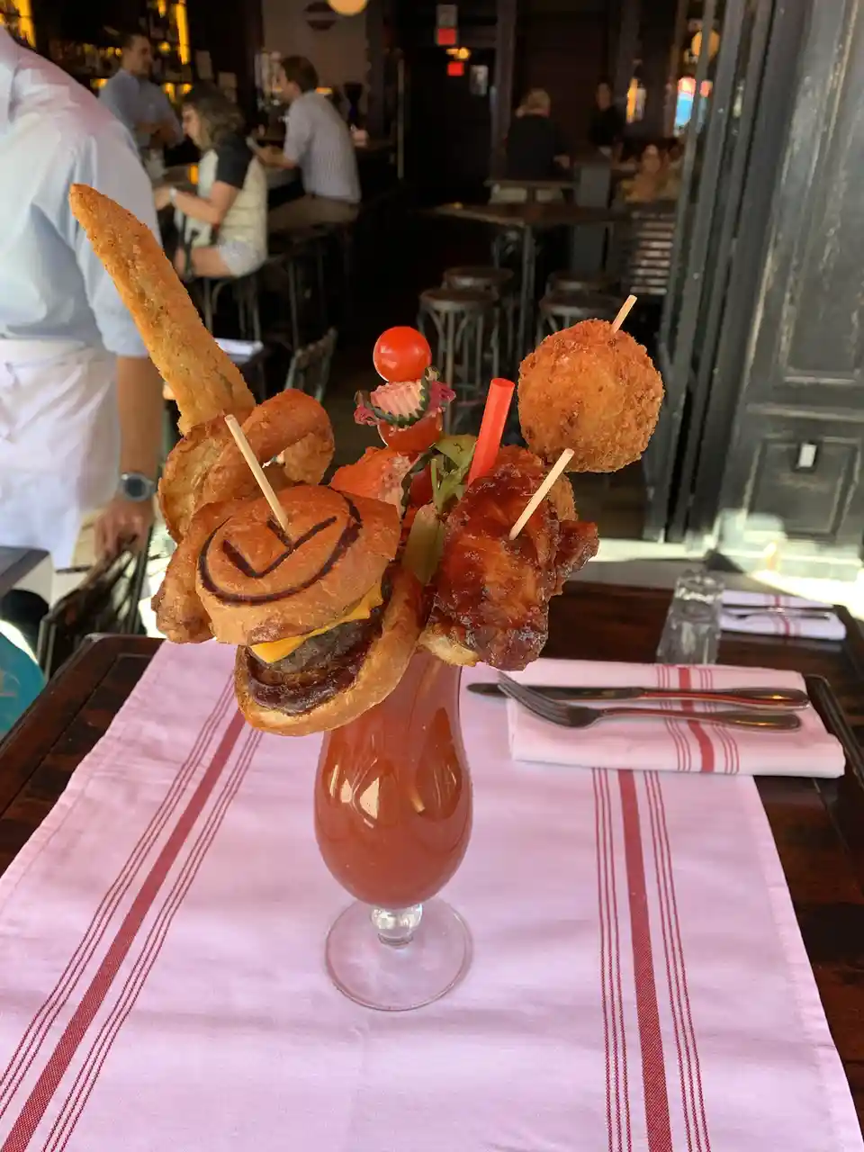 A tall, ornate cocktail glass filled with a reddish beverage is topped with an elaborate, chaotic arrangement of fried foods…