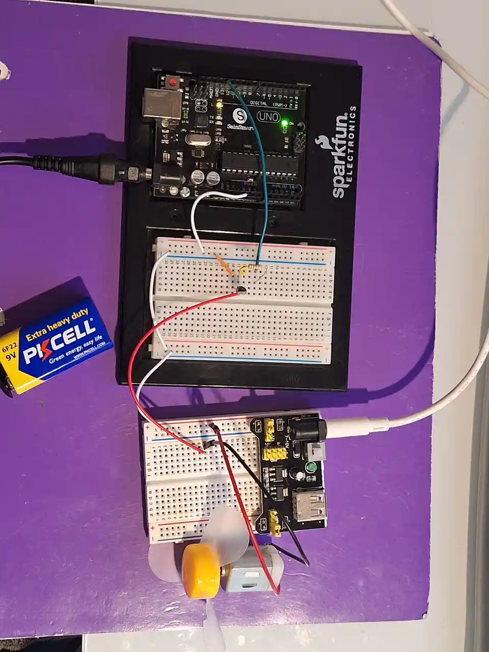 An overhead photo shows an electronics project on a purple surface, featuring an Arduino Uno board, two breadboards with…