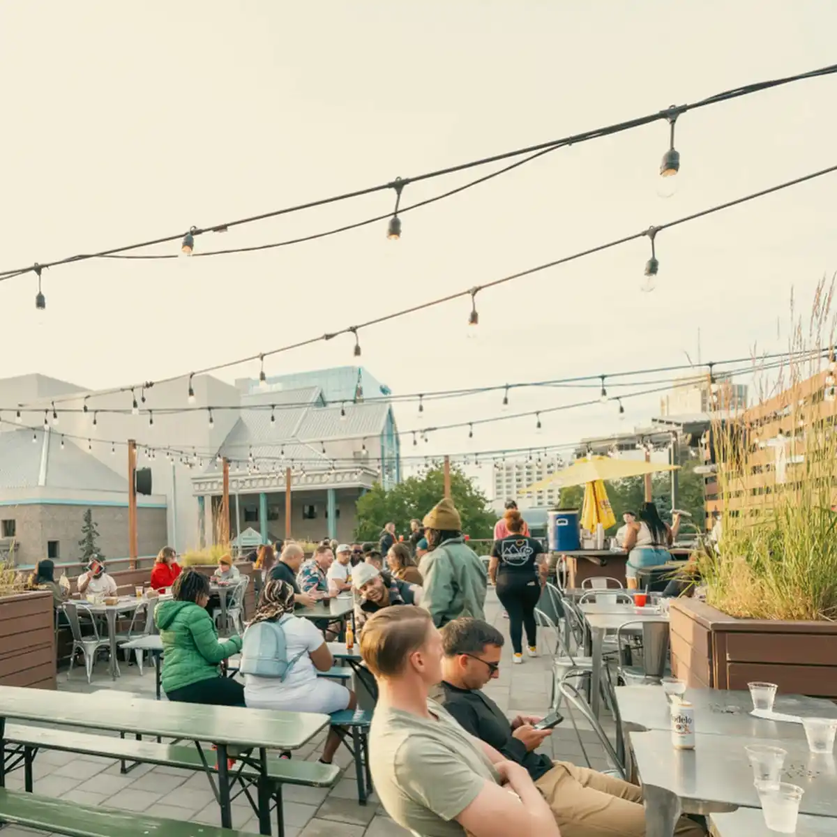 A crowded rooftop patio with string lights, people sitting at tables, and a man in the foreground looking away while another…