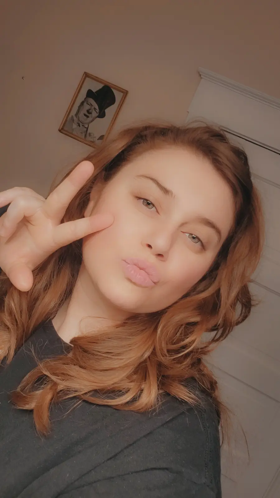 A young woman with long, wavy reddish-brown hair poses for a selfie, making a playful kissy face and holding up a peace sign…