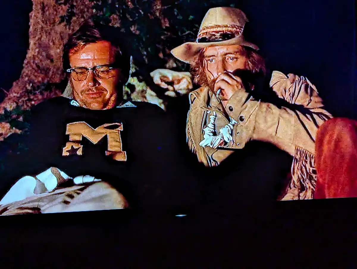 Two men are shown in a dimly lit, wooded setting. The man on the left wears glasses and a black shirt with a large gold 'M'…