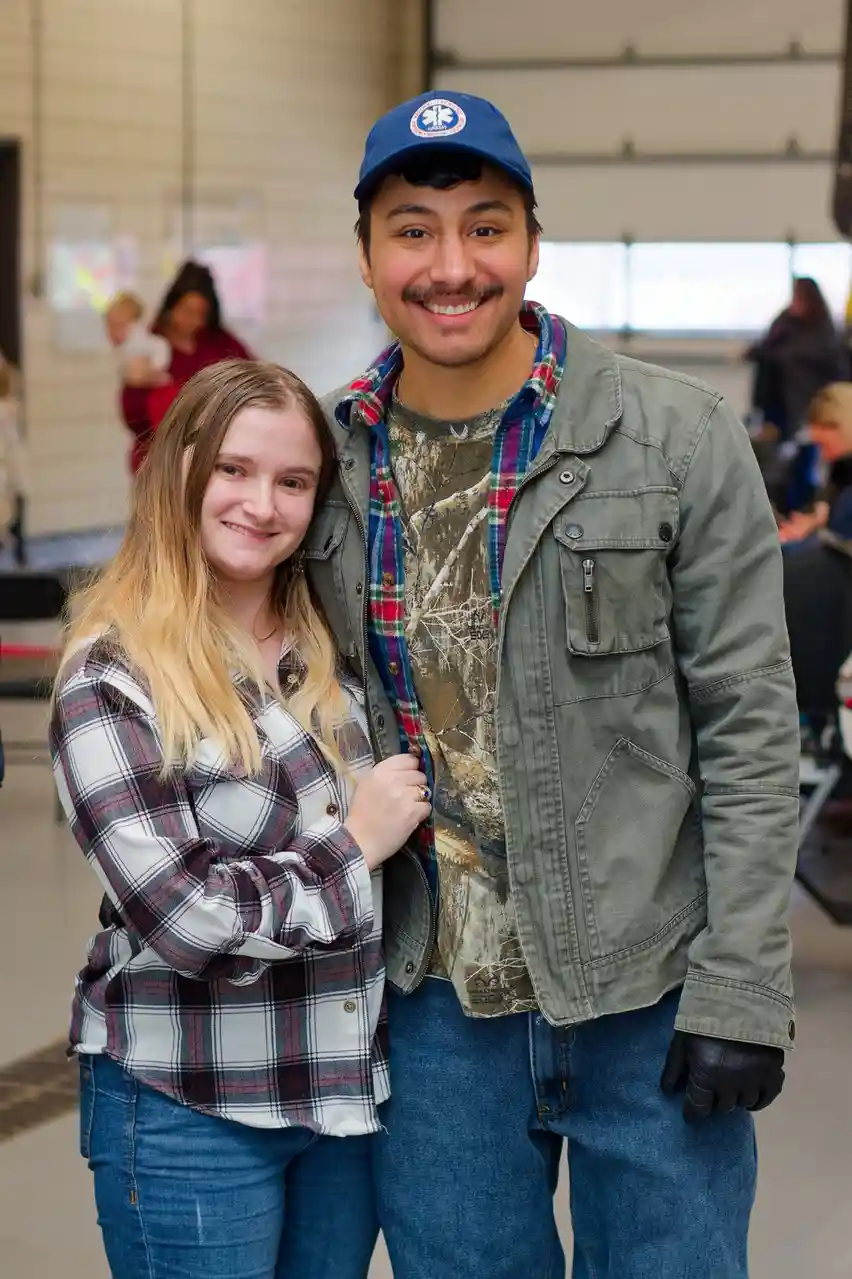 A young man and woman are posing for a photo together indoors. The man wears a blue cap, a camouflage t-shirt under a plaid…