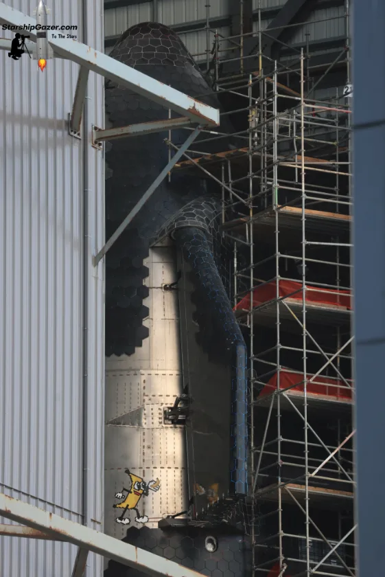 A large, hexagonal-patterned rocket or spacecraft structure is partially covered in scaffolding, with a cartoon image of…