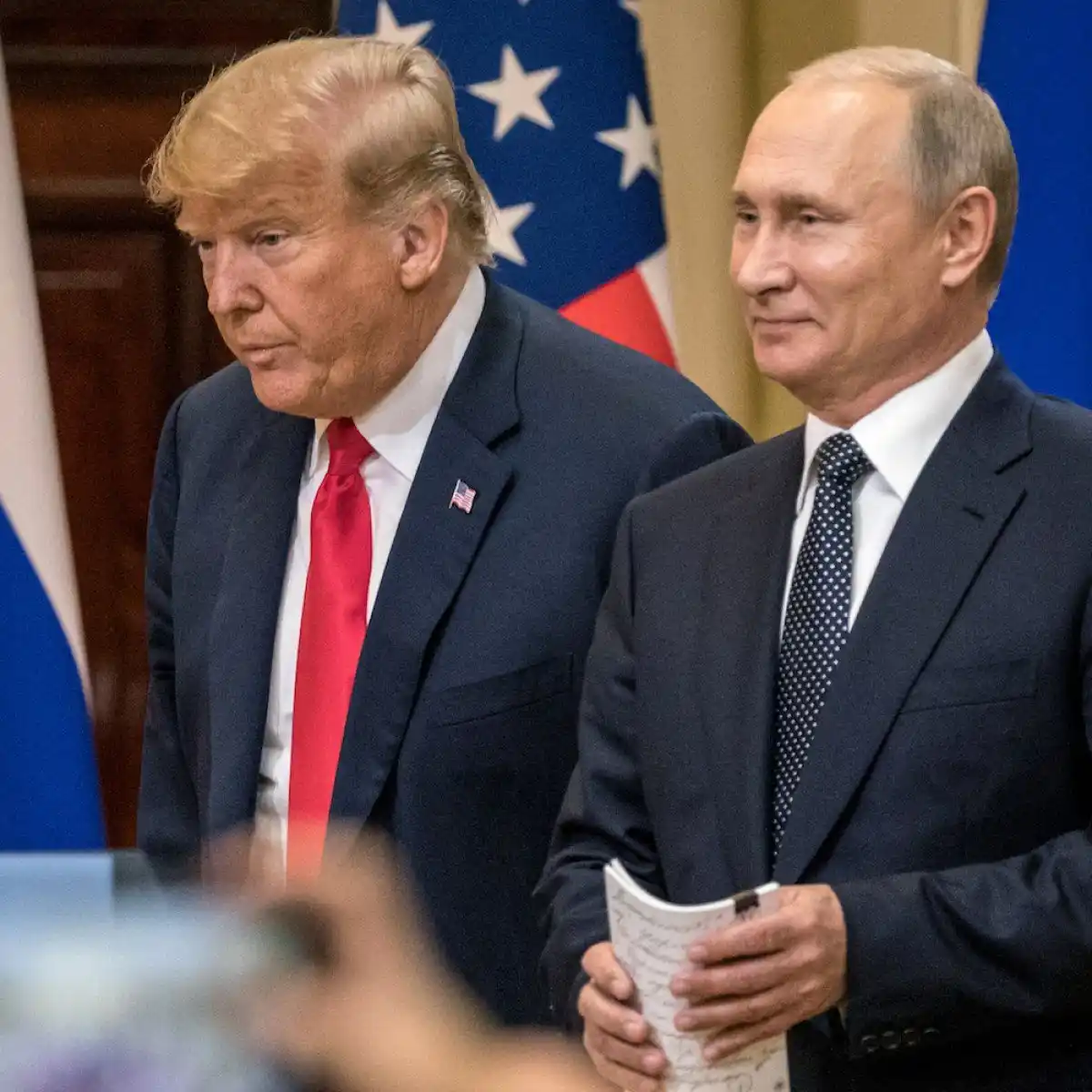 A photo of Donald Trump and Vladimir Putin standing side by side at a formal event, with American and Russian flags behind…