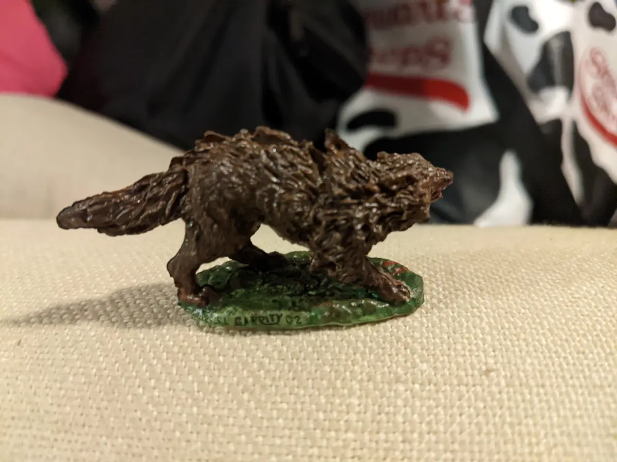 A small, brown, textured figurine of a wolf-like creature with exaggerated, shaggy fur stands on a green base. The figurine…