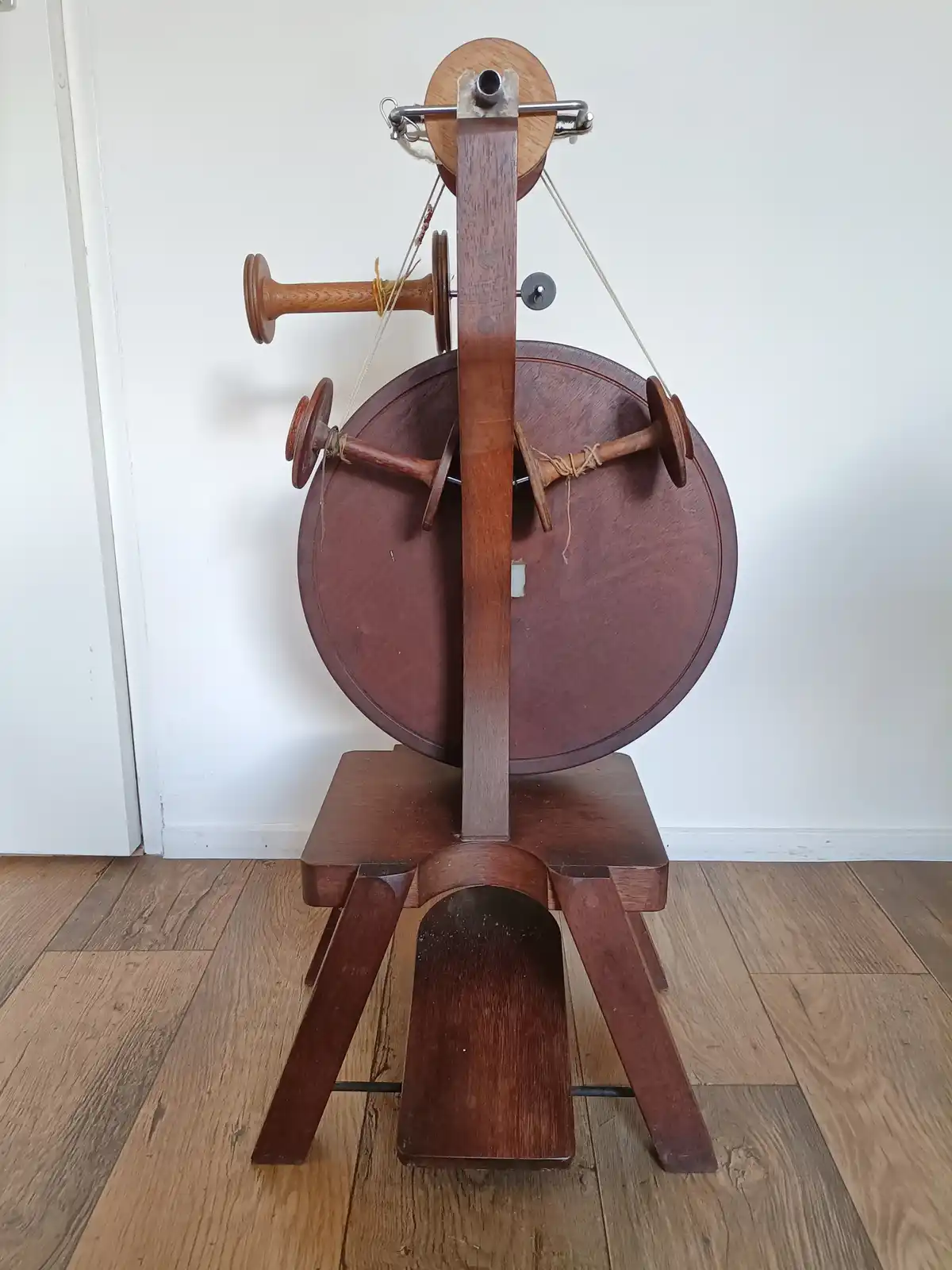 A wooden spinning wheel with multiple spindles and threads is shown standing on a wooden floor against a white wall. The…