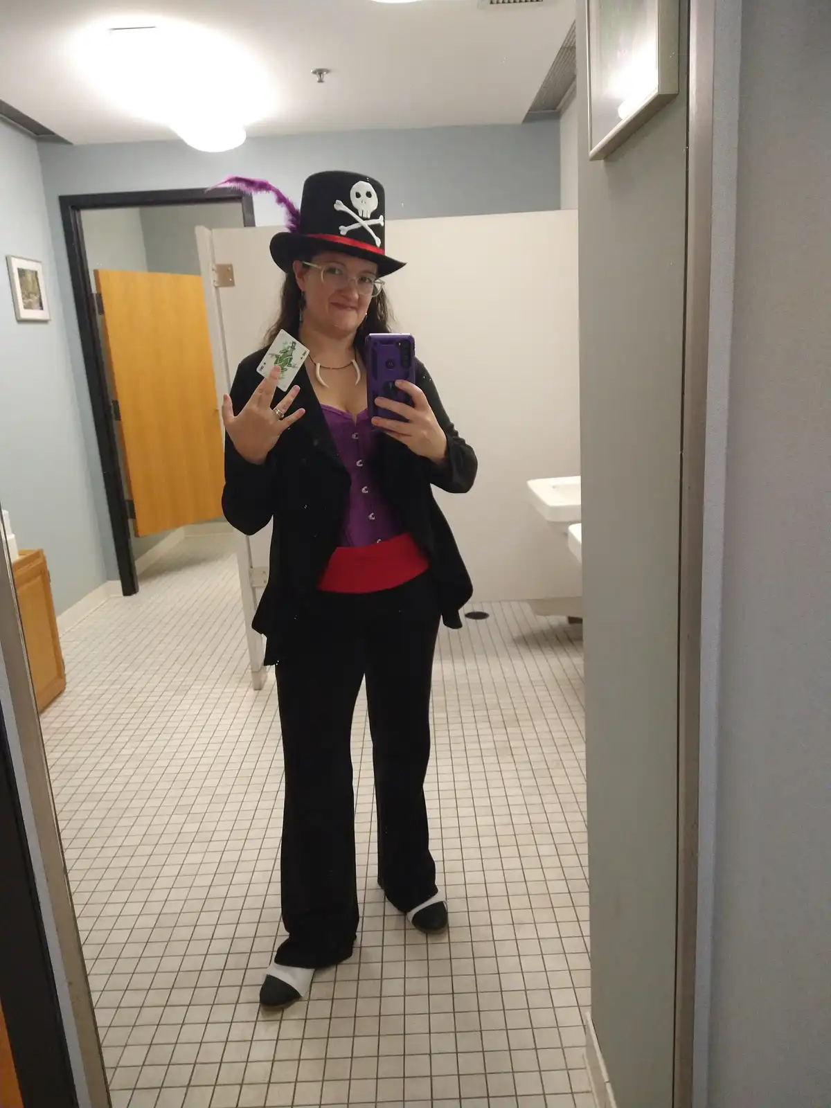 A person in a bathroom mirror selfie wearing a pirate hat with a skull and crossbones and a purple feather, a purple corset…