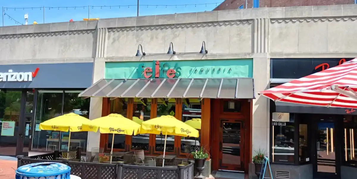 An outdoor shot of a restaurant named 'feip's taqueria' with yellow umbrellas and a glass awning, flanked by a Verizon store…