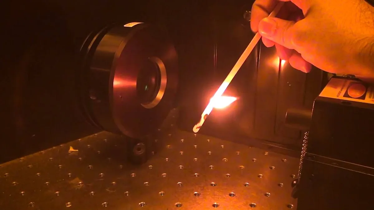 A hand holds a lit matchstick near a laser pointer device in a dark lab setting, with the laser beam visibly igniting the…