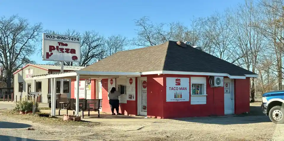 A red brick drive-thru-style food stand with a large sign reading 'The Pizza Place' and a smaller sign on the building that…
