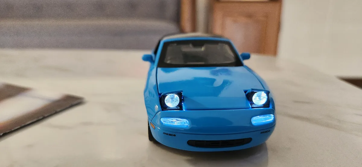A close-up photo of a blue Mazda Miata toy car with its headlights and fog lights illuminated, sitting on a light-colored…