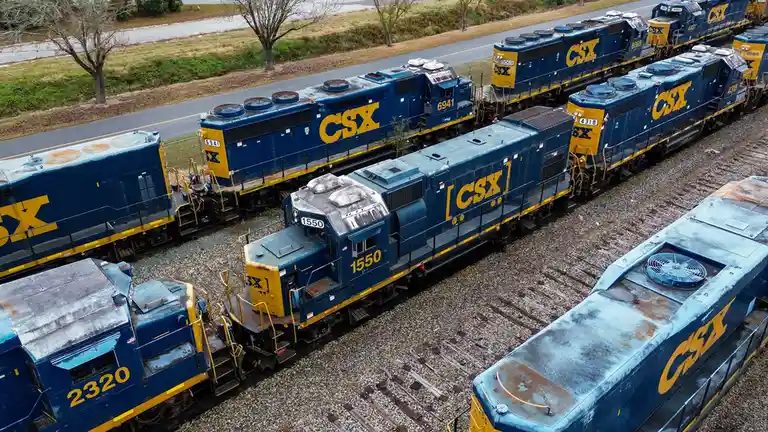 An aerial photo shows multiple blue and yellow CSX locomotives parked on parallel railroad tracks in a yard. The trains are…