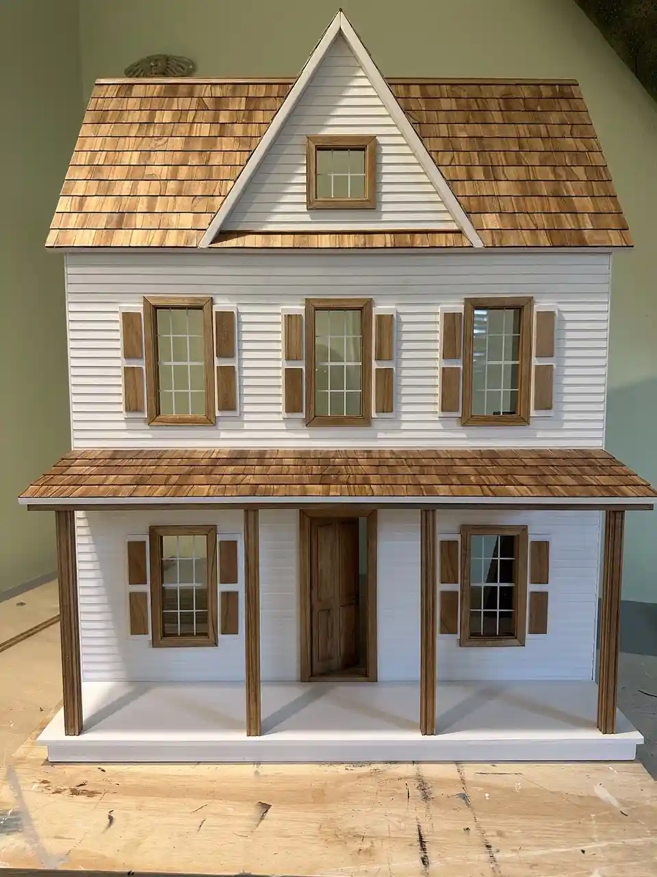 A detailed, two-story dollhouse with white siding, wooden shingle roof, and porch, sits on a wooden workbench. The text…