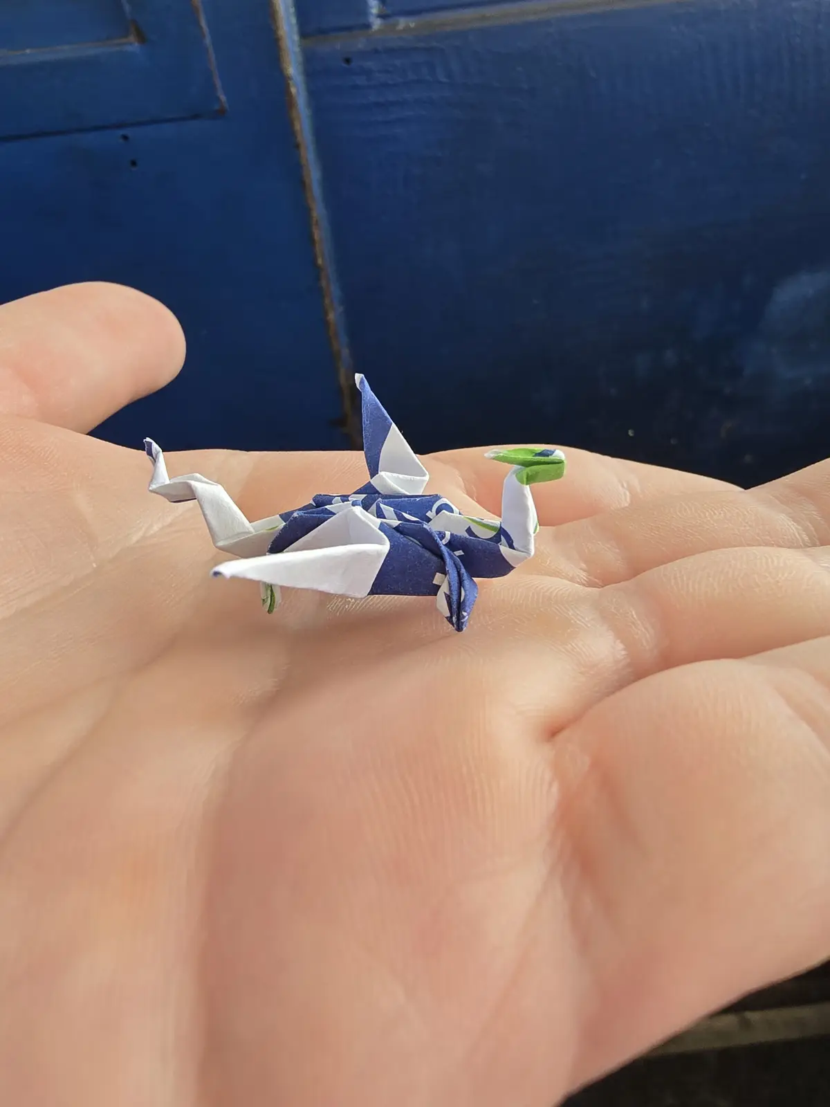 A tiny origami crane made from blue, white, and green patterned paper rests on an open palm, with a blue wooden surface in…