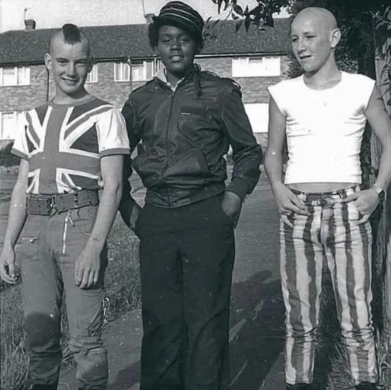 A black-and-white photo of three young men standing outdoors. The man on the left wears a Union Jack t-shirt and has a…