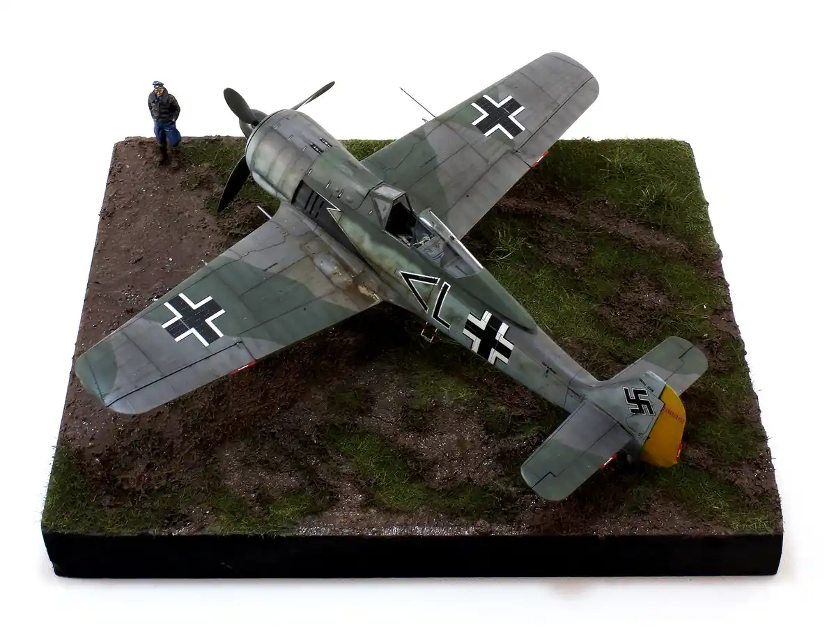 A detailed model of a German WWII fighter plane, likely a Messerschmitt Bf 109, is shown crashed on a diorama base with…