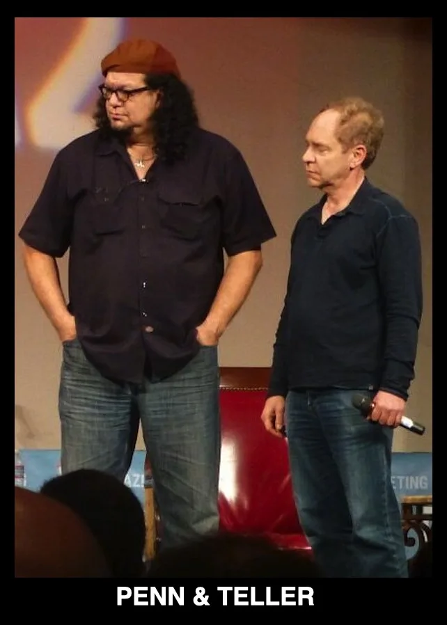 Two men stand on a stage, one with long curly hair, glasses, and a beret with hands in pockets, the other with short hair…