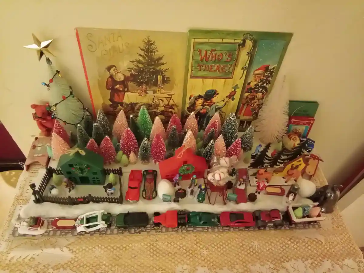 A cluttered Christmas display features vintage toys, miniature houses, bottlebrush trees, and classic Santa Claus imagery…