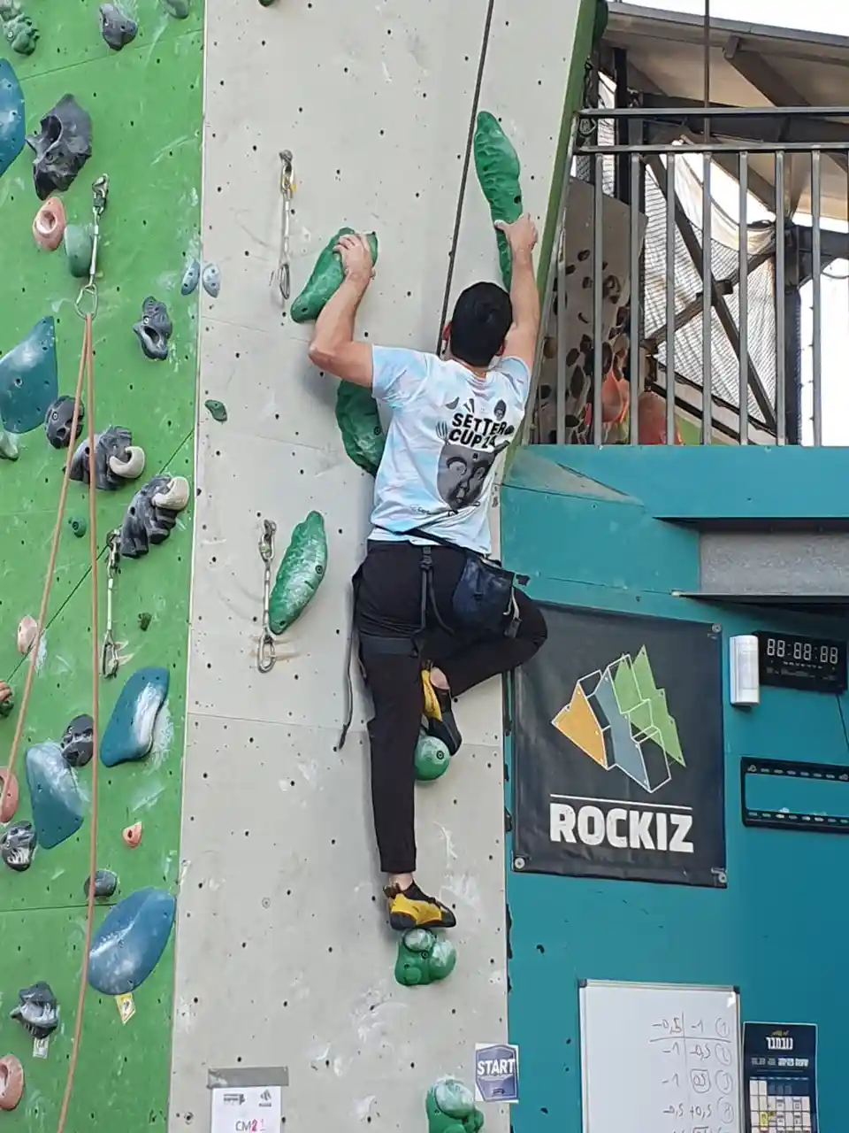 A male rock climber in a light blue 'Setter Cup 24' t-shirt is scaling an indoor climbing wall, gripping green holds with…