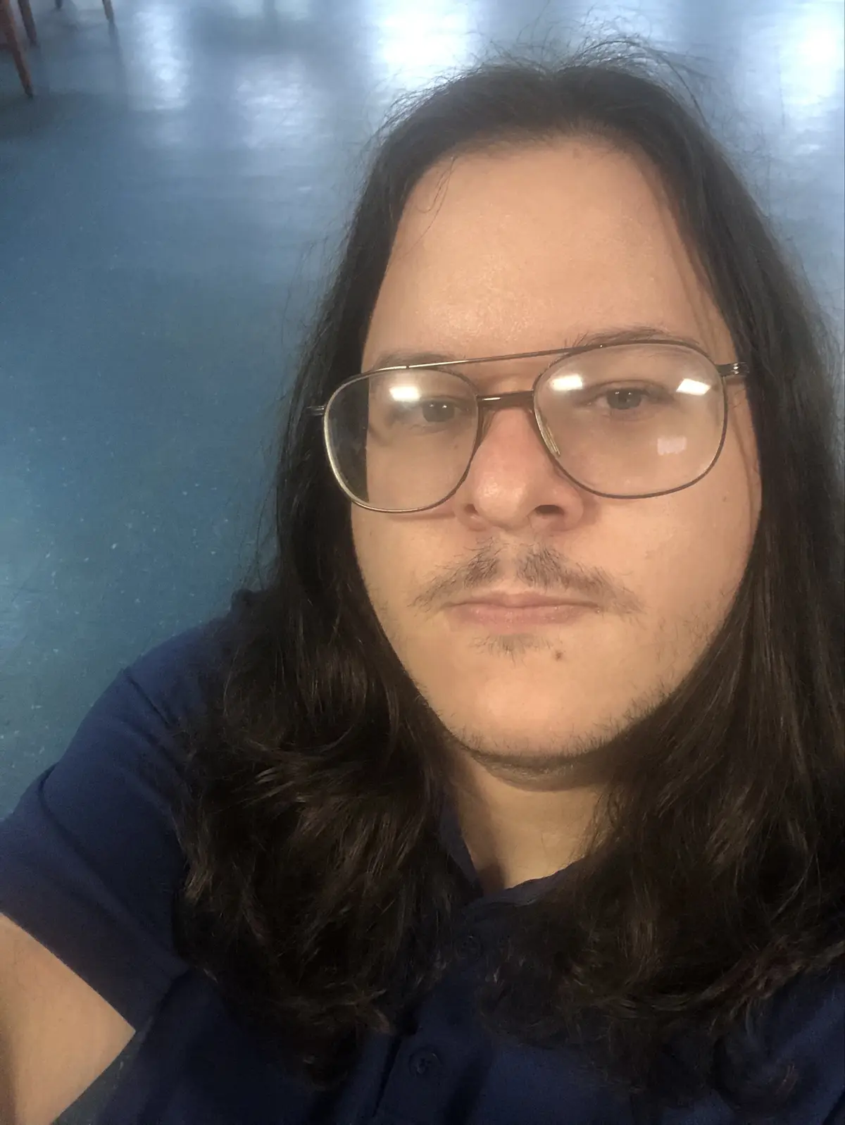 A person with long dark hair, wearing large rectangular glasses and a dark blue polo shirt, stares directly at the camera…