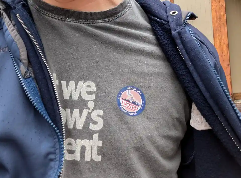 A close-up photo shows a person wearing a grey t-shirt under an unzipped blue jacket, with a circular 'I VOTED' sticker…