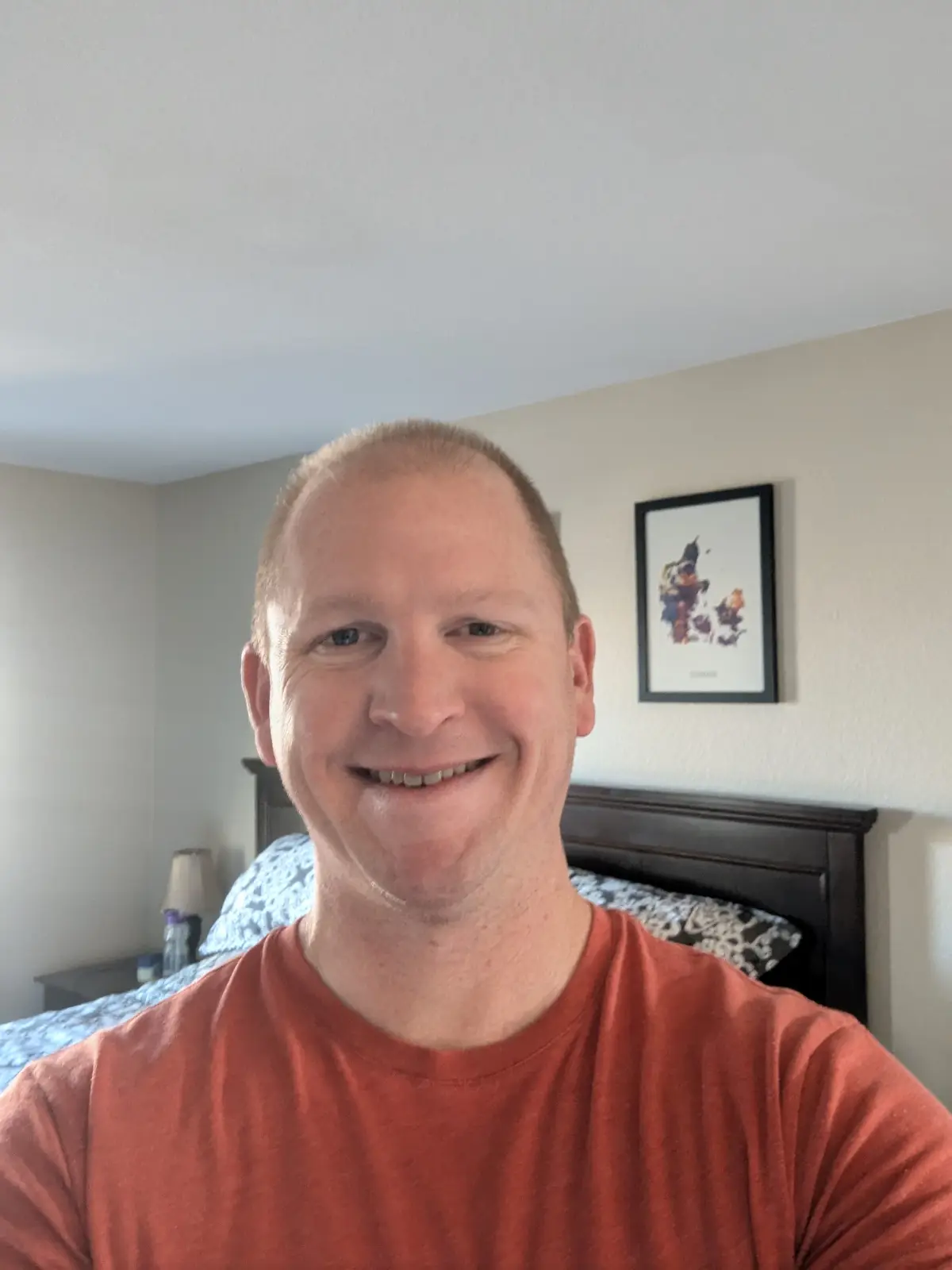A man with short blond hair and a wide, toothy smile takes a selfie in his bedroom, wearing a rust-colored t-shirt. Behind…