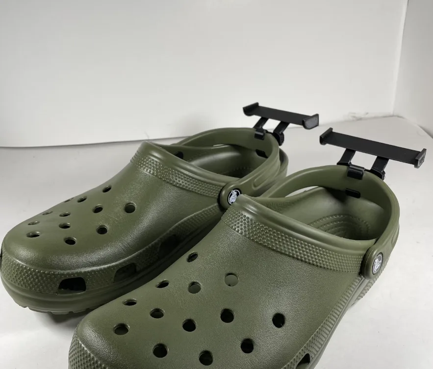 A pair of olive green Crocs clogs with black plastic racing spoilers attached to the back of each shoe, set against a plain…