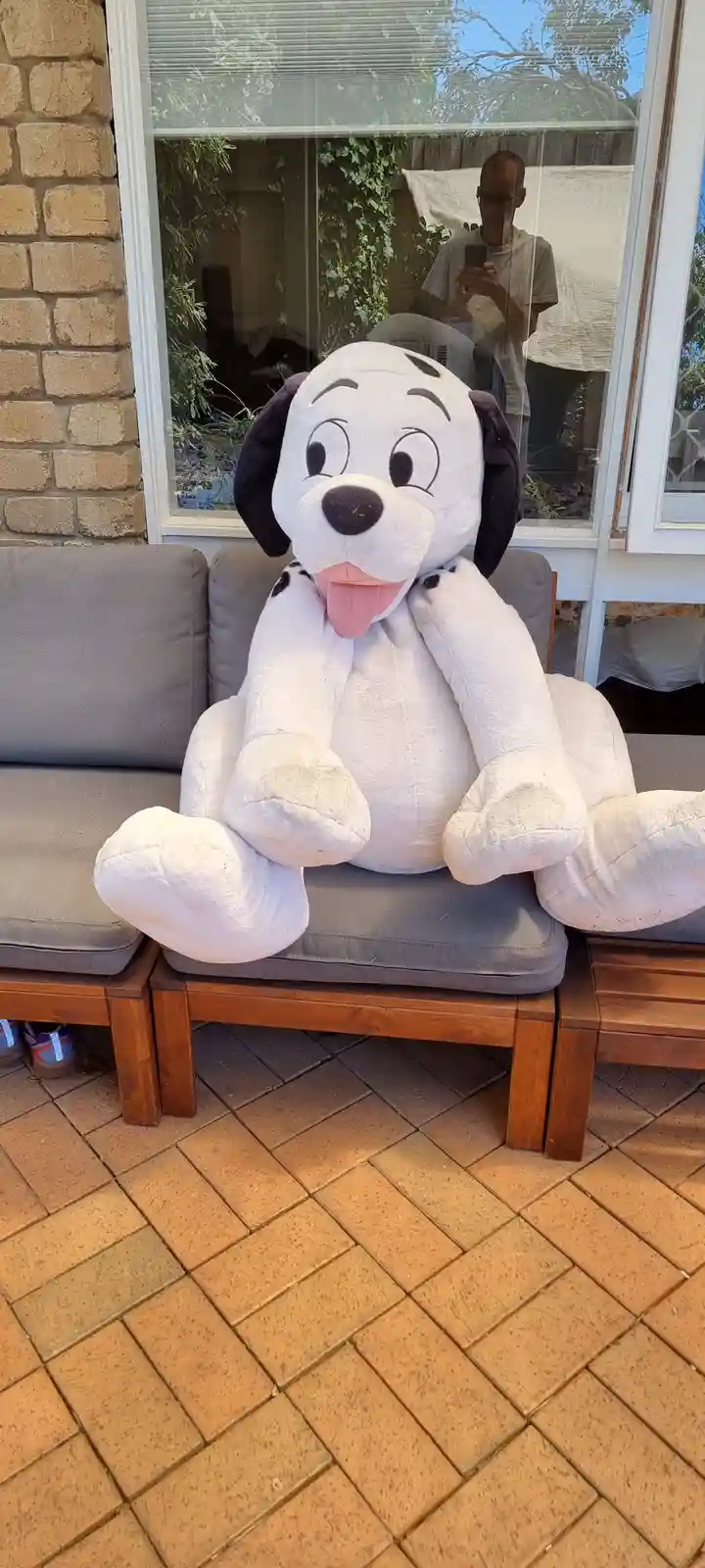 A giant, white plush Dalmatian dog with black spots and a pink tongue sits on a gray outdoor couch. In the reflection of the…