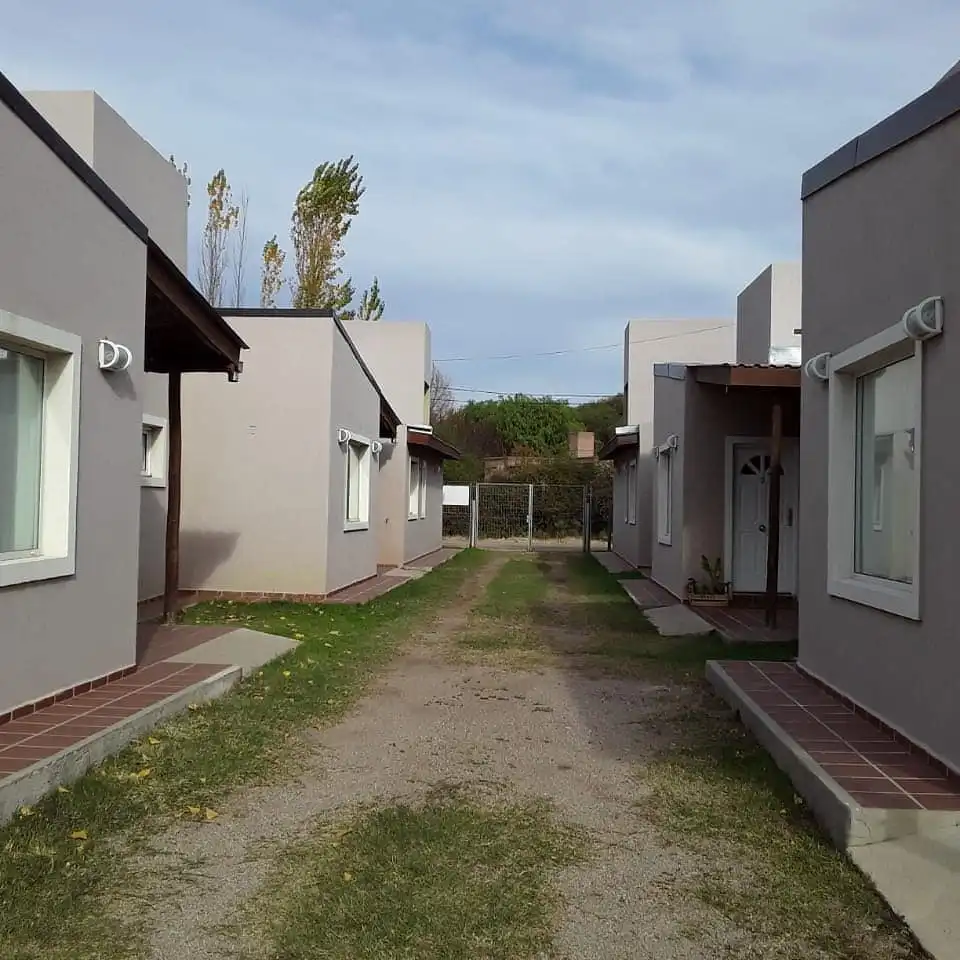 A narrow dirt path runs between two rows of identical modern, beige-colored single-story houses with white-trimmed windows…