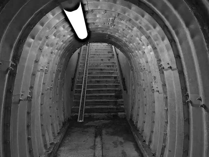 A black-and-white photo shows a long, narrow metal tunnel with a steep staircase ascending into the distance. The tunnel…