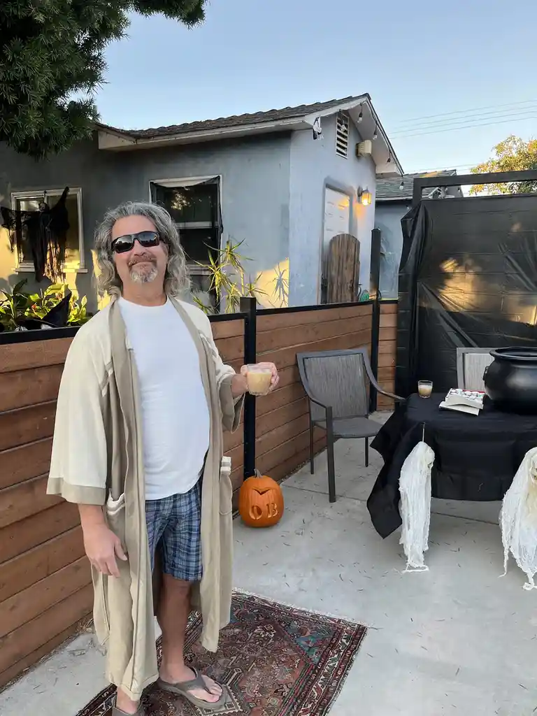 A man with long gray hair and sunglasses stands in a backyard wearing a robe and plaid shorts, holding a drink. Behind him…