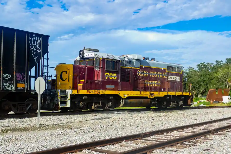 A maroon and yellow Ohio Central System locomotive, numbered 705 and 2114, sits on train tracks next to a graffiti-covered…