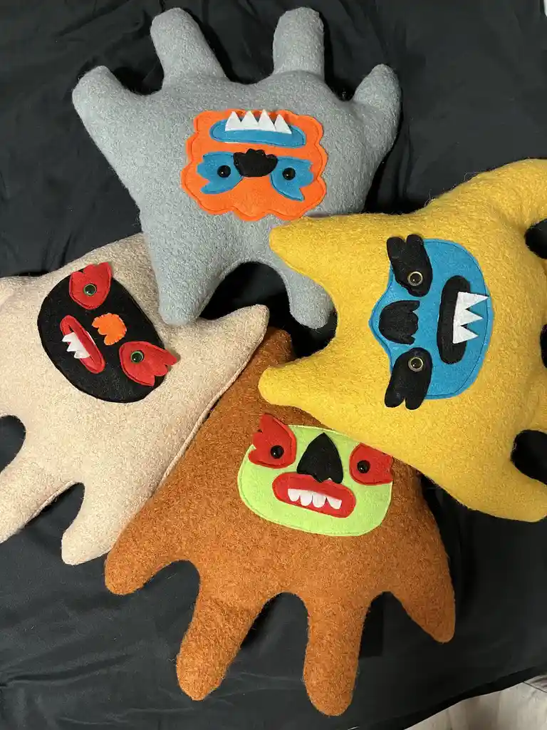 Four handmade, felt monster plushies shaped like hands, each with a colorful, stitched-on face featuring eyes, mouths, and…