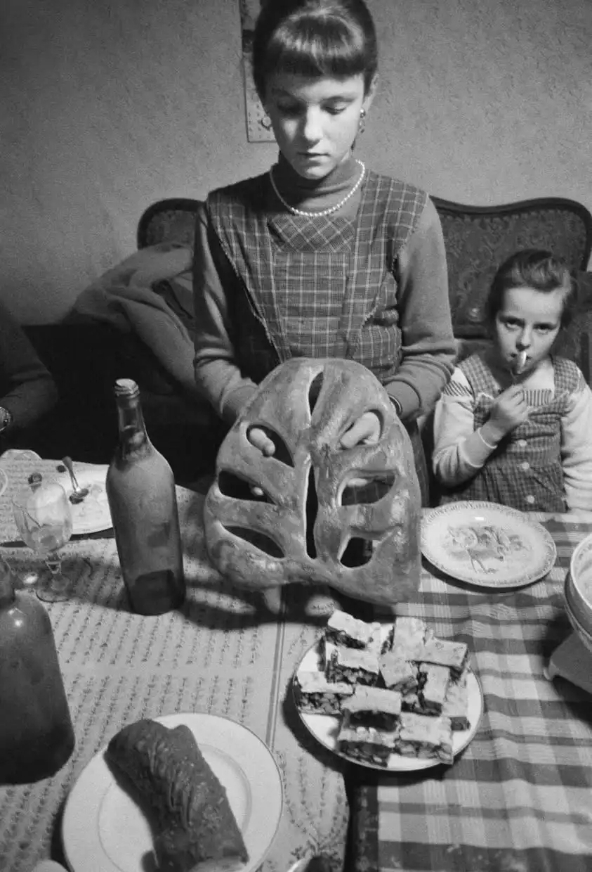 A black-and-white photo shows a young girl at a dinner table holding a large, carved bread loaf shaped like a human face…