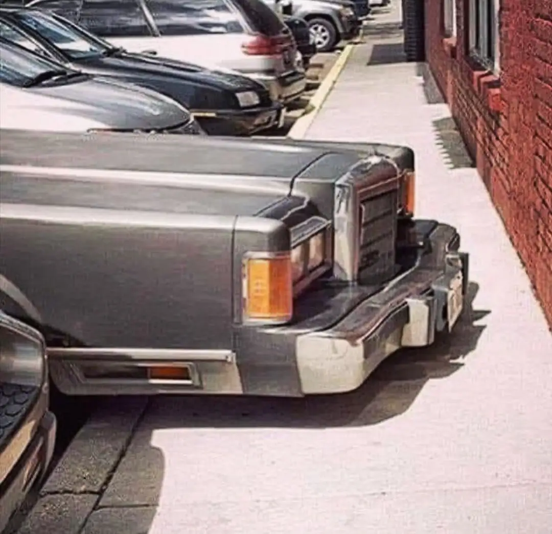 A silver, boxy, older model car is parked backwards in a narrow street parking spot, its rear bumper and tail lights facing…